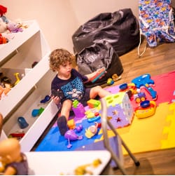 Criança repleta de brinquedos, sentada ao lado deles em um tatame de espuma, segurando vários brinquedos nas mãos.